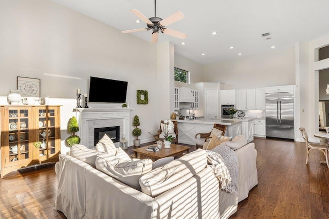 living area with ceiling fan, high vaulted ceiling, dark wood-style flooring, baseboards, and a glass covered fireplace