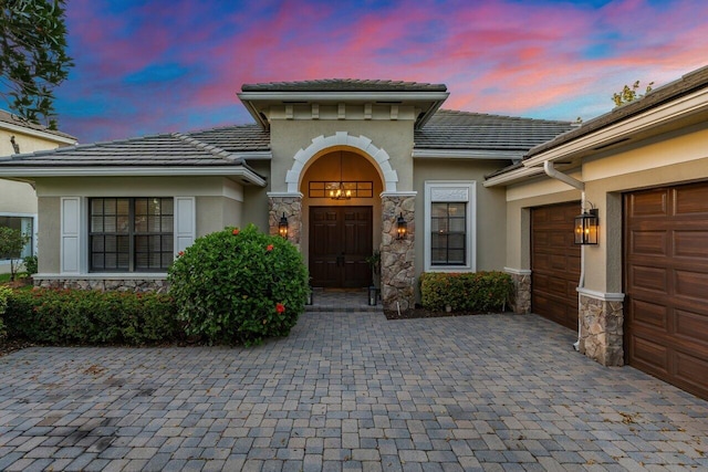 exterior entry at dusk with decorative driveway, a tile roof, stucco siding, an attached garage, and stone siding