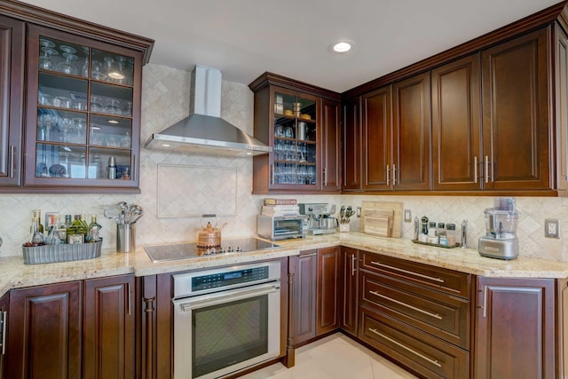 kitchen with tasteful backsplash, light stone counters, black electric stovetop, wall chimney range hood, and stainless steel oven