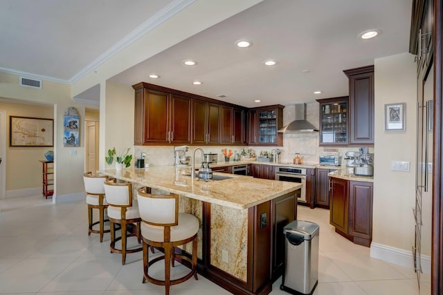 kitchen featuring visible vents, stainless steel oven, a sink, wall chimney range hood, and a peninsula