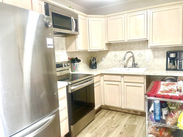 kitchen with tile counters, appliances with stainless steel finishes, a sink, light wood-type flooring, and backsplash