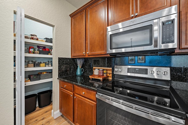 kitchen featuring a textured wall, light wood-style flooring, appliances with stainless steel finishes, dark stone countertops, and backsplash