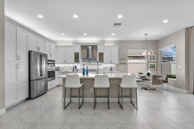 kitchen with wall chimney exhaust hood, a kitchen island with sink, stainless steel appliances, light countertops, and backsplash