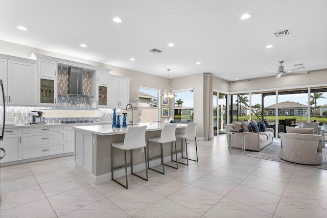 kitchen with visible vents, an island with sink, wall chimney exhaust hood, open floor plan, and light countertops