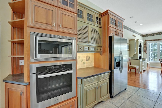 kitchen with tasteful backsplash, dark countertops, appliances with stainless steel finishes, glass insert cabinets, and light tile patterned flooring