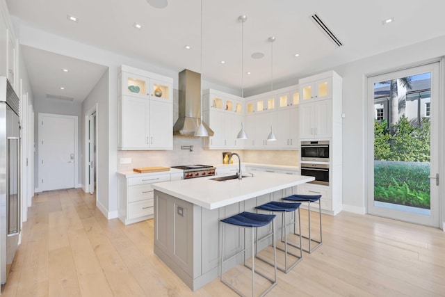 kitchen with a kitchen island with sink, a sink, wall chimney range hood, tasteful backsplash, and light wood-type flooring