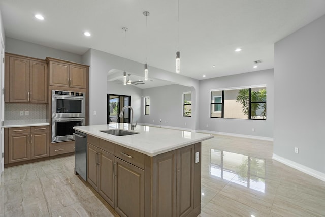 kitchen with a center island with sink, a ceiling fan, a sink, double oven, and backsplash