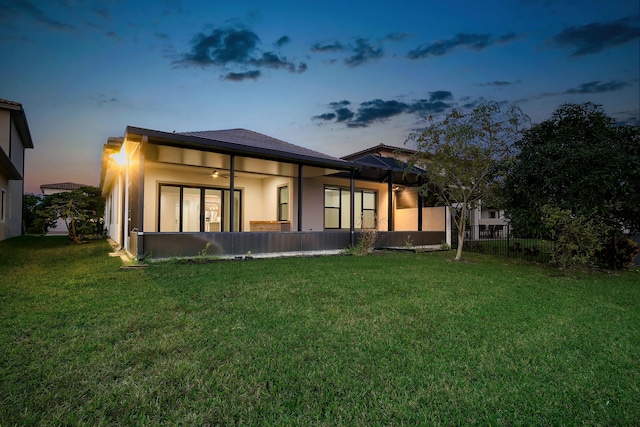 back of property at dusk with a lawn, fence, a sunroom, and stucco siding