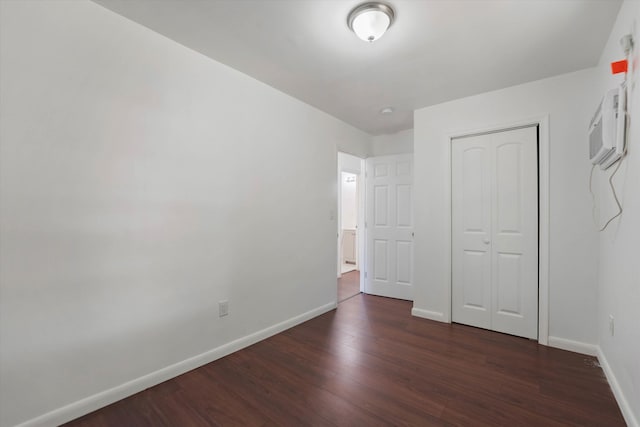 unfurnished bedroom with a wall unit AC, a closet, baseboards, and dark wood-type flooring