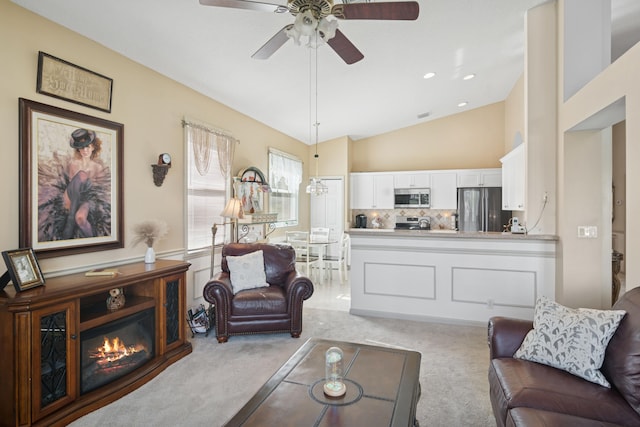 living area featuring lofted ceiling, ceiling fan, a glass covered fireplace, and light colored carpet