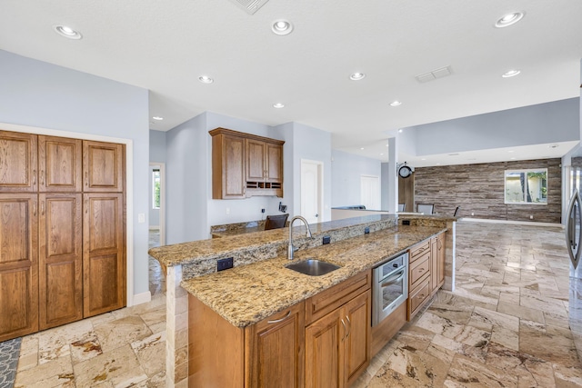 kitchen featuring a center island with sink, light stone counters, brown cabinets, a sink, and recessed lighting