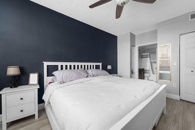 bedroom featuring an accent wall, light wood-style floors, a textured ceiling, ensuite bath, and baseboards