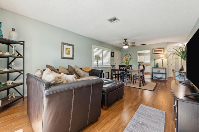 living room with a textured ceiling, light wood-style flooring, visible vents, and a ceiling fan