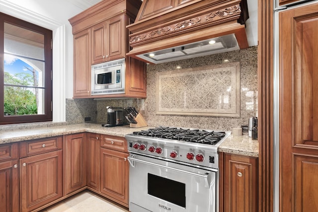 kitchen featuring brown cabinets, custom range hood, decorative backsplash, appliances with stainless steel finishes, and light stone countertops