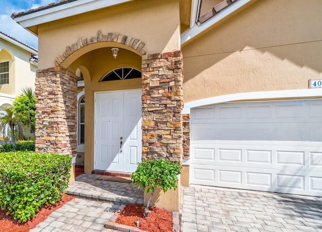 view of exterior entry featuring decorative driveway, stone siding, an attached garage, and stucco siding