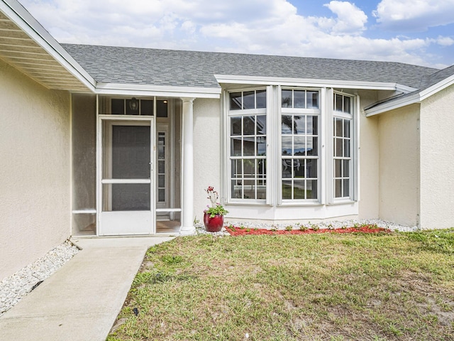 view of exterior entry featuring roof with shingles, a lawn, and stucco siding