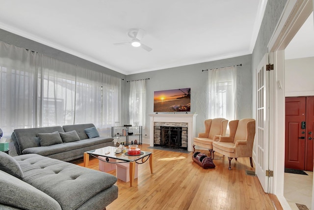 living room featuring ceiling fan, a stone fireplace, light wood-type flooring, and crown molding