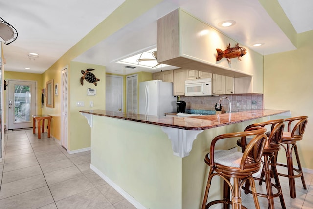 kitchen featuring light tile patterned floors, a peninsula, white appliances, decorative backsplash, and dark stone countertops