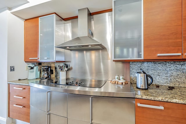 kitchen with glass insert cabinets, light stone counters, wall chimney range hood, black electric stovetop, and tasteful backsplash