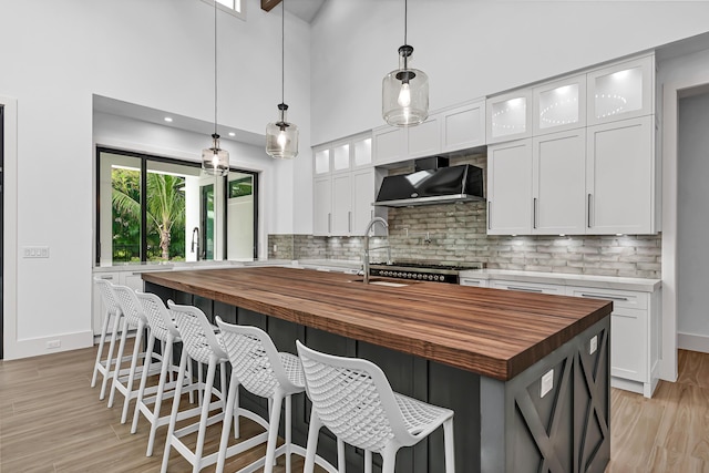 kitchen with butcher block countertops, white cabinets, wall chimney range hood, light wood finished floors, and tasteful backsplash