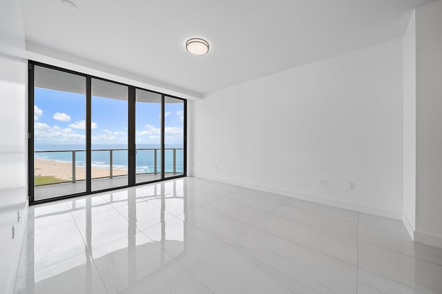 tiled empty room with a wall of windows, a water view, a view of the beach, and baseboards