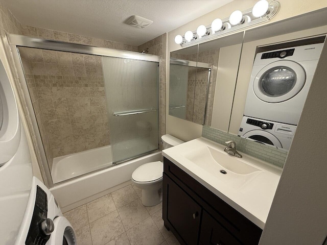 full bathroom featuring bath / shower combo with glass door, stacked washer / dryer, toilet, tile patterned floors, and a textured ceiling