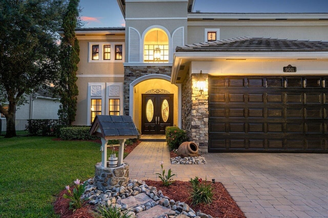 exterior entry at dusk with french doors, stucco siding, a lawn, a garage, and stone siding