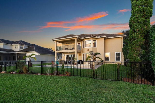 back of property at dusk with a balcony, fence private yard, stucco siding, and a yard