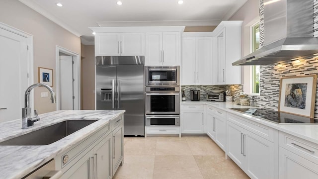 kitchen with built in appliances, a sink, white cabinetry, backsplash, and wall chimney exhaust hood