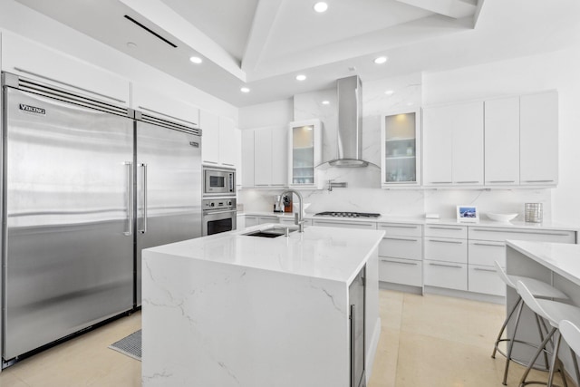 kitchen featuring wall chimney range hood, white cabinets, built in appliances, and a sink