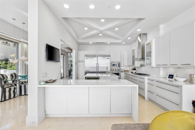 kitchen featuring wall chimney range hood, built in appliances, light countertops, a wealth of natural light, and a peninsula