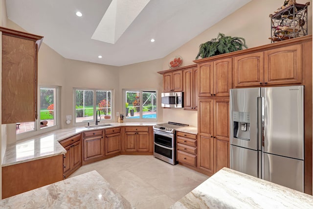 kitchen with brown cabinetry, lofted ceiling with skylight, light stone counters, appliances with stainless steel finishes, and a sink