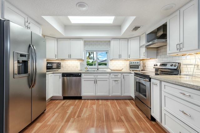 kitchen featuring a raised ceiling, appliances with stainless steel finishes, light wood-type flooring, wall chimney range hood, and a sink