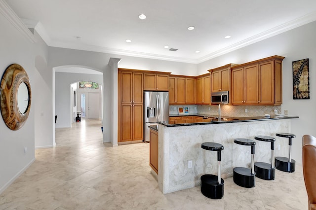 kitchen with arched walkways, brown cabinetry, appliances with stainless steel finishes, a peninsula, and a kitchen bar