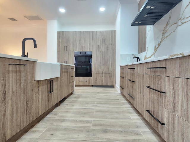 kitchen with light wood finished floors, modern cabinets, multiple ovens, ventilation hood, and light brown cabinetry