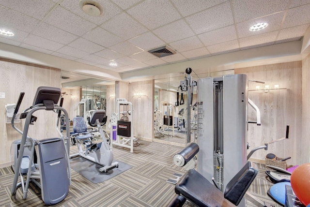 exercise room with carpet floors, visible vents, wooden walls, and a drop ceiling