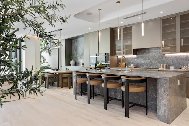 kitchen with decorative backsplash, modern cabinets, a kitchen island with sink, light wood-type flooring, and stone counters