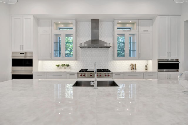 kitchen with light stone counters, double oven, white cabinets, a sink, and wall chimney exhaust hood