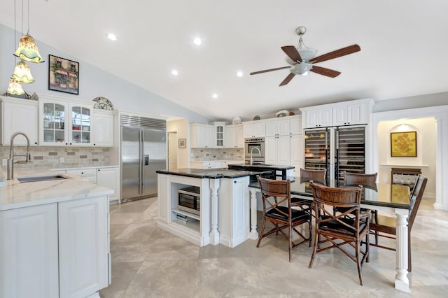 kitchen with white cabinets, an island with sink, dark stone countertops, built in appliances, and a sink