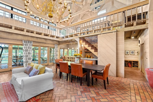 dining space featuring brick floor, wooden walls, a notable chandelier, a high ceiling, and french doors