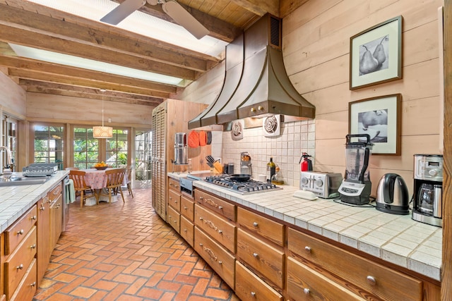 kitchen with tile counters, custom range hood, brick floor, wood walls, and a sink