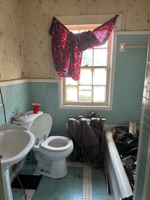 bathroom featuring tile walls, toilet, a bathing tub, wainscoting, and wallpapered walls