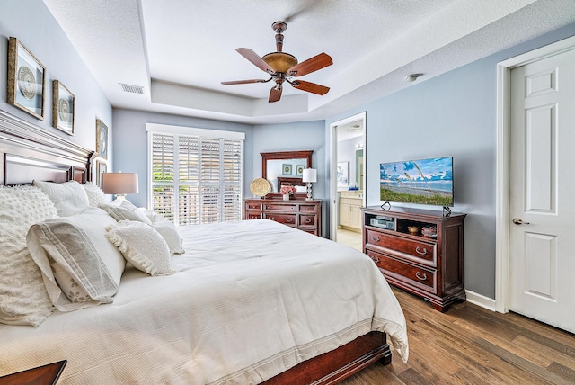 bedroom featuring a tray ceiling, visible vents, ensuite bathroom, wood finished floors, and baseboards