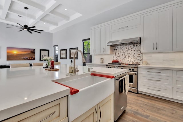 kitchen with coffered ceiling, wall chimney exhaust hood, stainless steel appliances, light wood-style floors, and backsplash