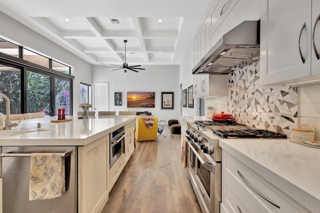 kitchen with light wood finished floors, backsplash, appliances with stainless steel finishes, wall chimney range hood, and coffered ceiling