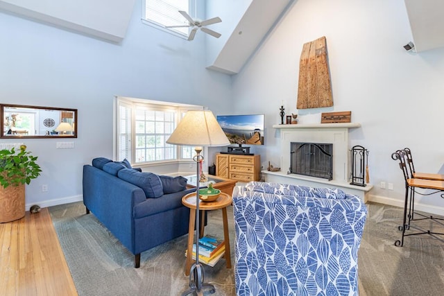 living area featuring a fireplace with raised hearth, high vaulted ceiling, ceiling fan, wood finished floors, and baseboards