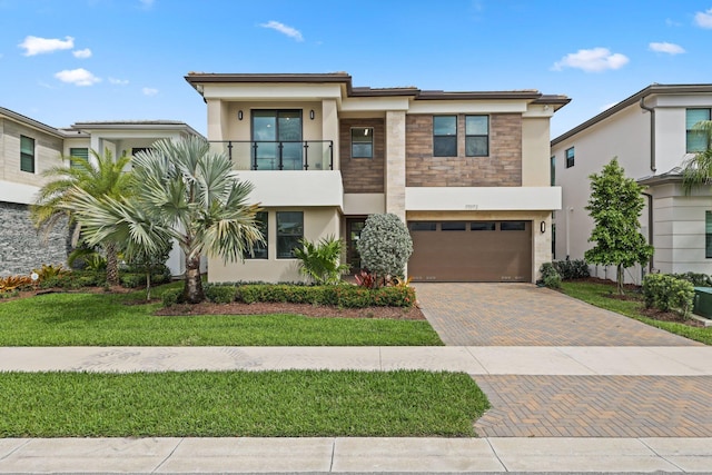 view of front facade featuring decorative driveway, stucco siding, an attached garage, a balcony, and stone siding