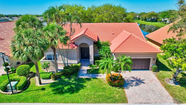 mediterranean / spanish-style house with an attached garage, a tiled roof, decorative driveway, stucco siding, and a front yard