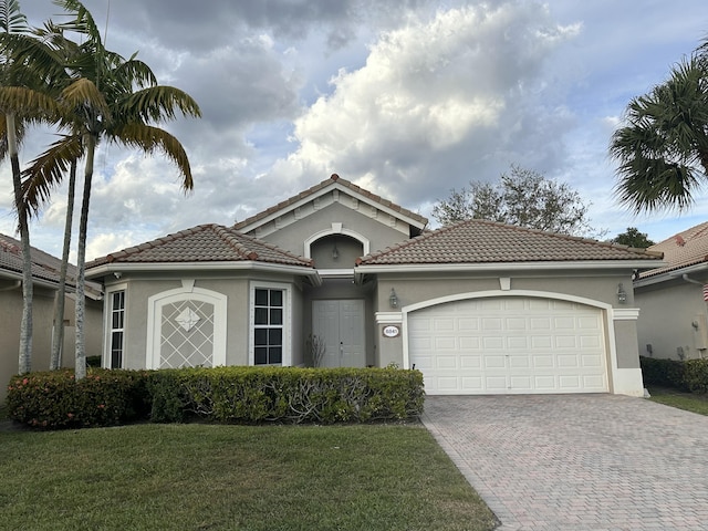 mediterranean / spanish home featuring decorative driveway, stucco siding, a garage, a tiled roof, and a front lawn