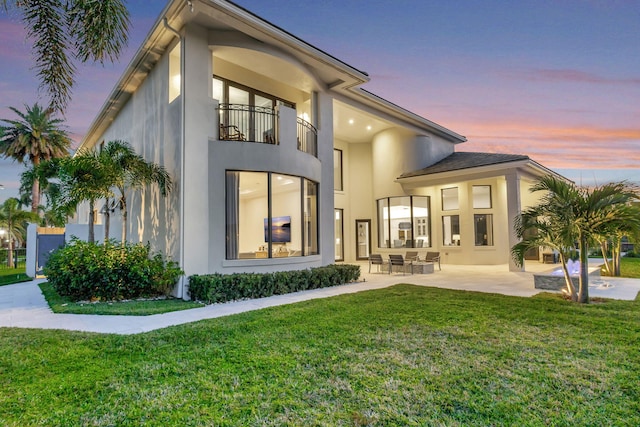 back of property at dusk featuring stucco siding, a yard, a balcony, and a patio